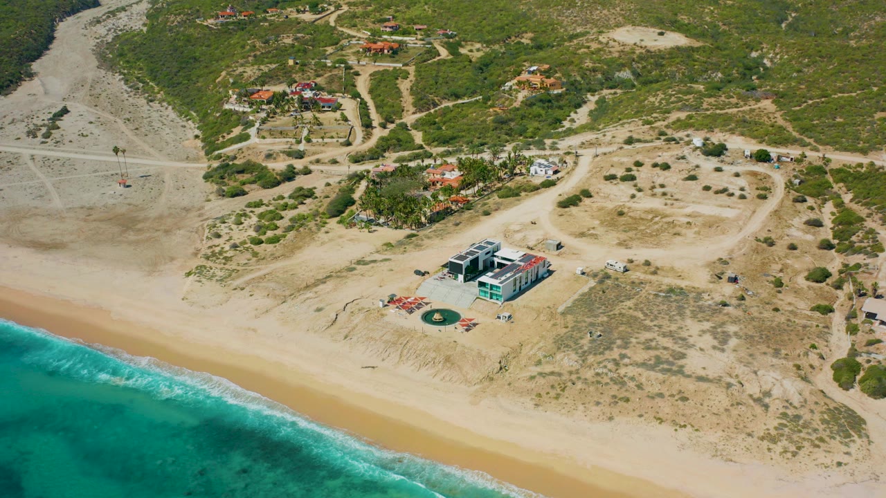 Aerial frame over El Cardoncito, Baja California Sur, with turquoise water at lower left, pale sand, a beachfront house near center, inland tracks, scattered homes, and no visible people.