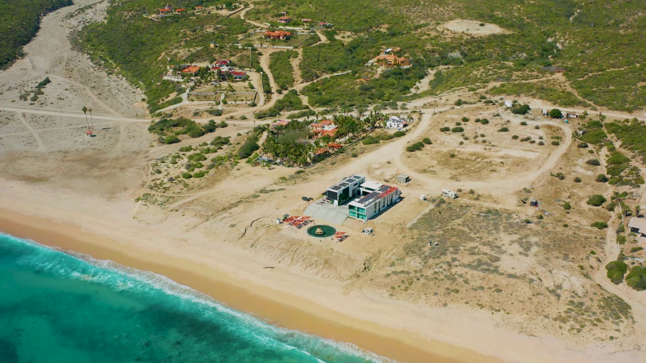 Drone-height view in El Cardoncito with clear blue-green water edging the sand, a modern beachfront structure below center, dry lots, winding dirt tracks, scattered homes, and no visible people.