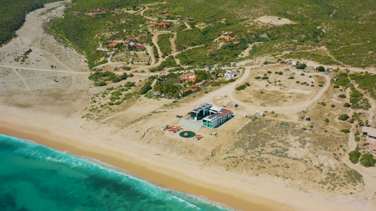 Aerial still from El Cardoncito showing turquoise surf in the lower left, a wide pale beach, a boxy beachfront building, dry scrubland, dirt access tracks, distant houses, and no visible people.