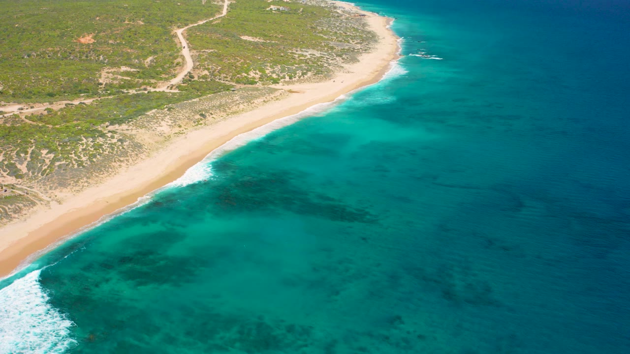 High aerial view of El Cardoncito coastline with pale sand, turquoise shallows, darker reef-like water patches, low green vegetation, sandy tracks inland, and no visible people.