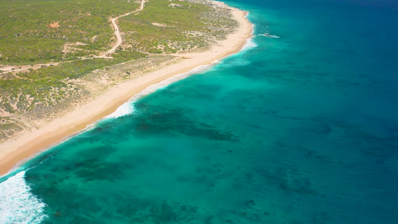 Aerial frame over El Cardoncito showing the beach curving along clear blue water, small white waves on the lower left shore, scrub-covered land above, and no visible people.