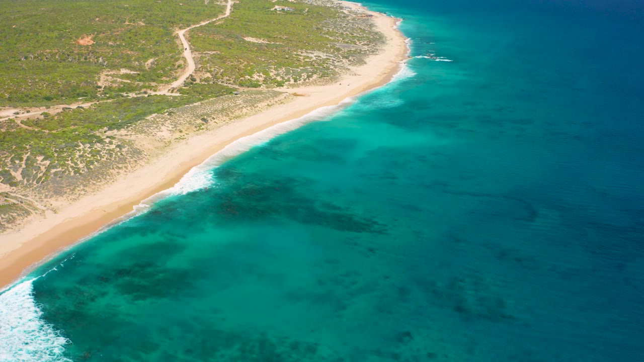 Aerial view of El Cardoncito with sandy coast at the left, bright turquoise shallows fading into deeper blue sea, faint underwater shadows, inland dirt tracks, and no visible people.