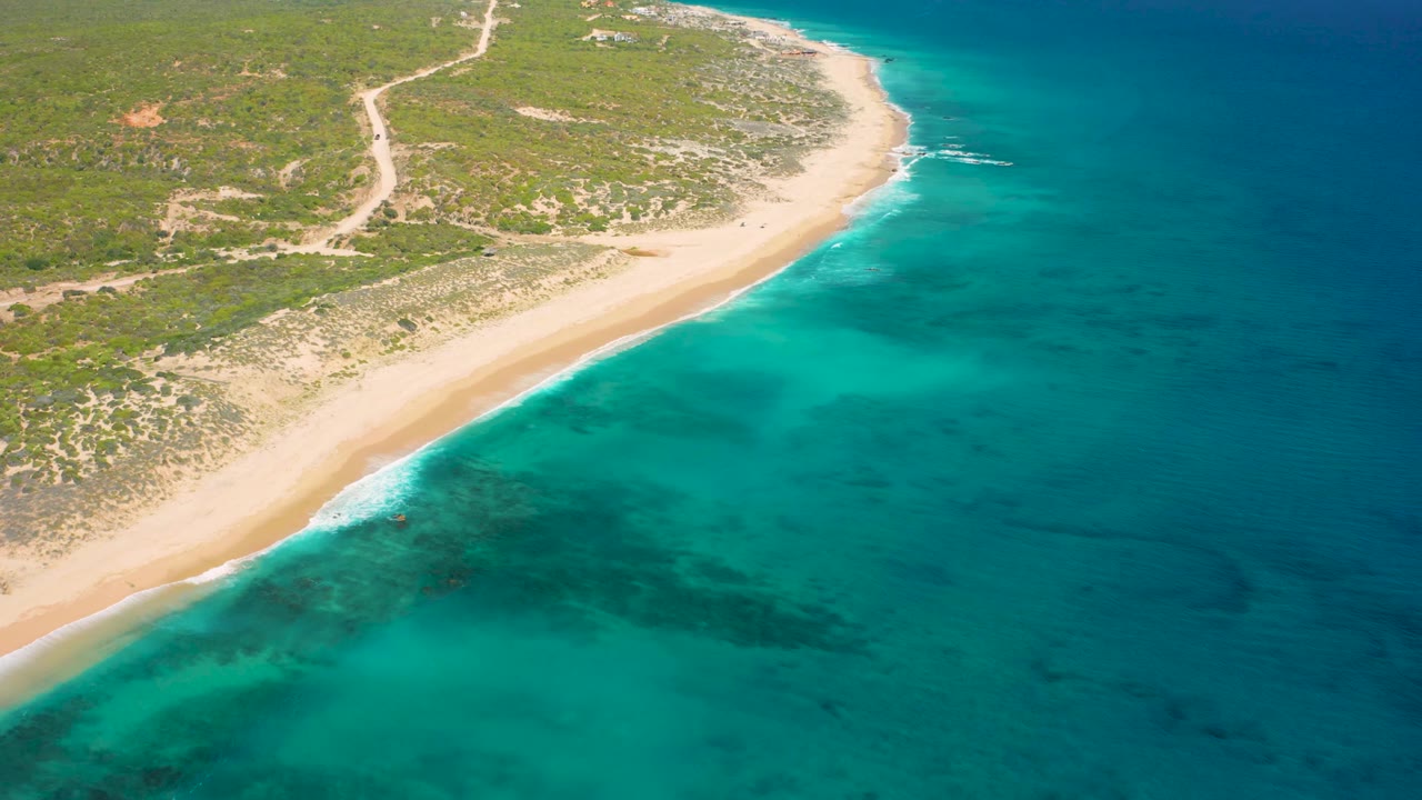 High oblique aerial frame of El Cardoncito with open turquoise water in the foreground, a narrow sandy beach along the left, scrubland behind it, and no visible people.
