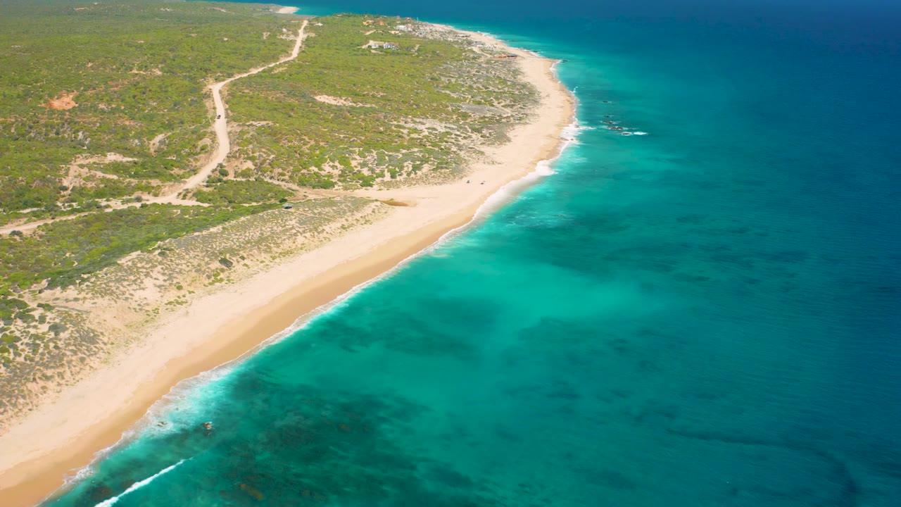 High-angle El Cardoncito coastline view with the beach arc slightly farther along the frame, bright shallow water near shore, deeper blue water offshore, and no visible people.