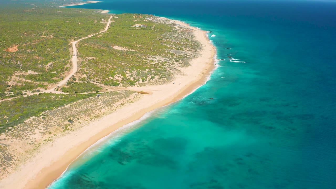 Aerial view over El Cardoncito with the sandy shoreline running diagonally through the frame, blue-green water on the right, scrubland and a dirt track inland, and no visible people.