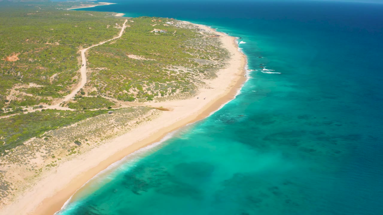 Aerial frame from El Cardoncito showing pale sand against turquoise shallows, darker blue sea farther out, inland brush, a visible dirt road, and no visible people.