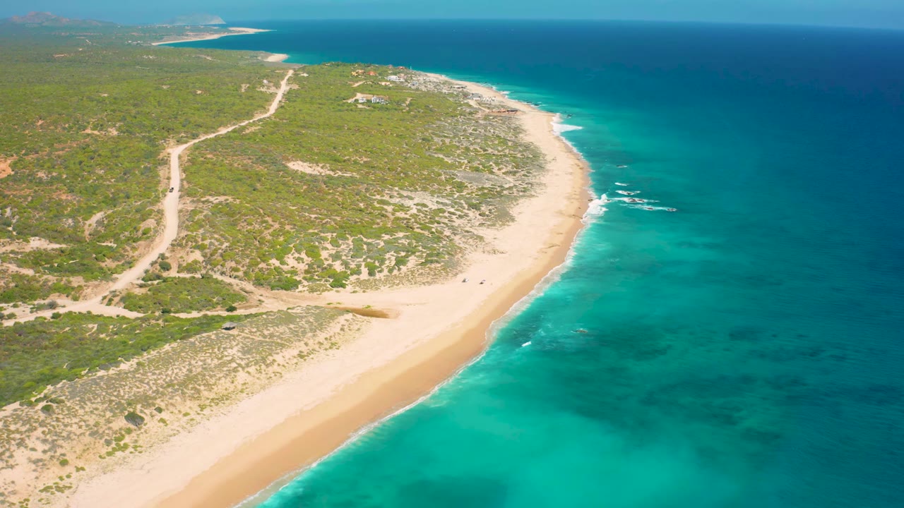 El Cardoncito coastline from above, with the beach curve slightly farther along the frame, blue-green water to the right, scrubland and road inland, and no visible people.