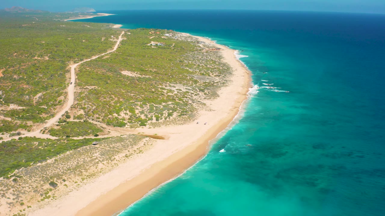 Aerial view of El Cardoncito with more open sea visible on the right, pale sand along the center, green vegetation inland, a winding dirt road, and no visible people.