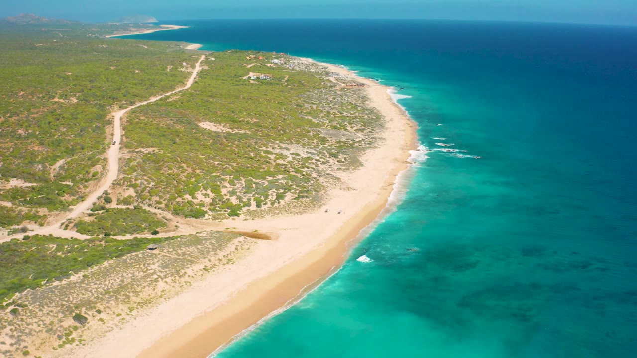 Overhead coastal frame from El Cardoncito showing the sandy shore angled across the lower half, turquoise surf along the edge, green scrubland inland, and no visible people.