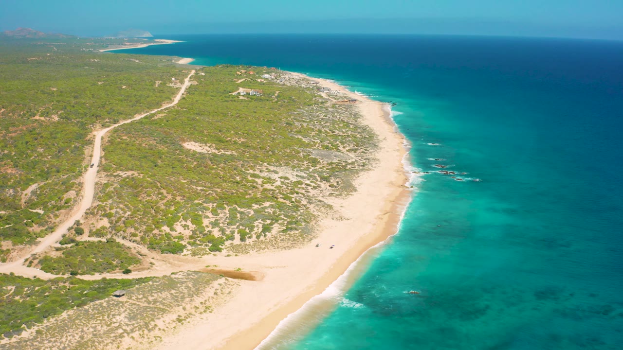 Wide aerial view in El Cardoncito, Baja California Sur, with scrub-covered land to the left, a pale beach running through the frame, calm turquoise water to the right, and no visible people.