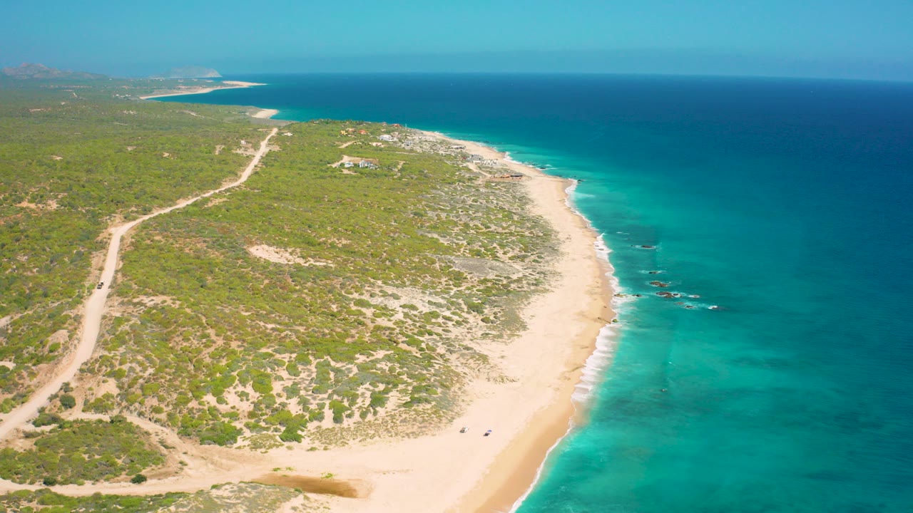 Aerial frame over El Cardoncito, Baja California Sur showing green coastal scrub, a pale beach, turquoise shallows, deeper blue sea, distant structures, and no visible people.