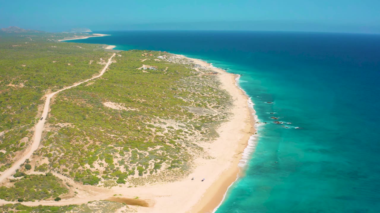 Aerial view at El Cardoncito with the dirt road more centered through the scrub, the beach running diagonally, small surf breaks offshore, distant structures, and no visible people.