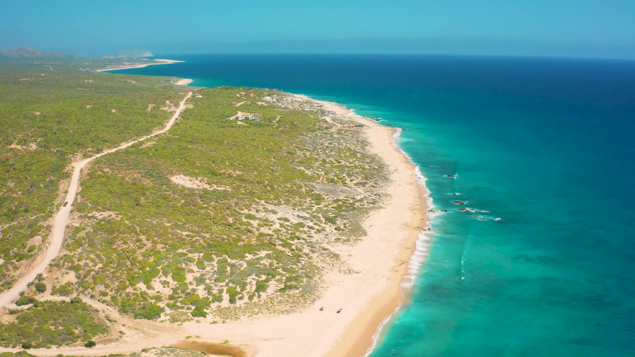 Elevated El Cardoncito frame showing the beach and shallows shifted slightly right, with scrubland and a pale road on the left, wave lines near offshore rocks, distant structures, and no visible people.