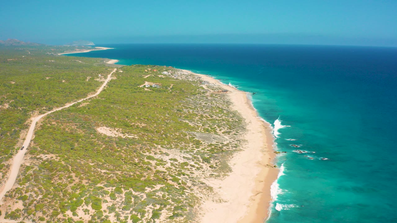 High aerial view over El Cardoncito, Baja California Sur, with scrub-covered land at left, a pale beach below, turquoise surf at right, and no visible people.