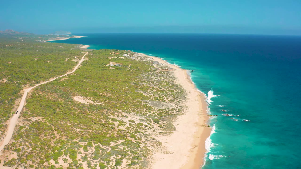 Aerial frame from El Cardoncito showing the beach curving through the center, low vegetation inland, bright shallows along the shore, and no visible people.