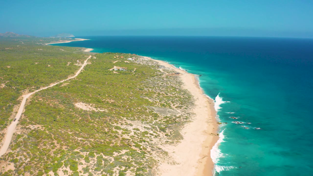 High coastal view in El Cardoncito with a dirt road through scrubland, pale sand along the water, a small rocky surf area offshore, and no visible people.