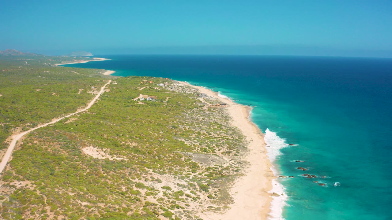 High aerial view over El Cardoncito, Baja California Sur, with turquoise water on the right, a pale beach below, low scrubland and dirt tracks inland, and no visible people.