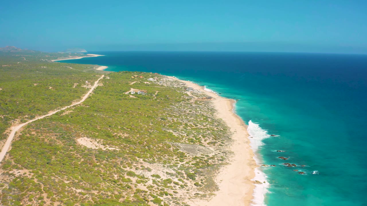 Drone-height view of El Cardoncito coastline with a diagonal sandy beach, bright turquoise shallows, darker ocean beyond, low vegetation, small inland structures, and no visible people.