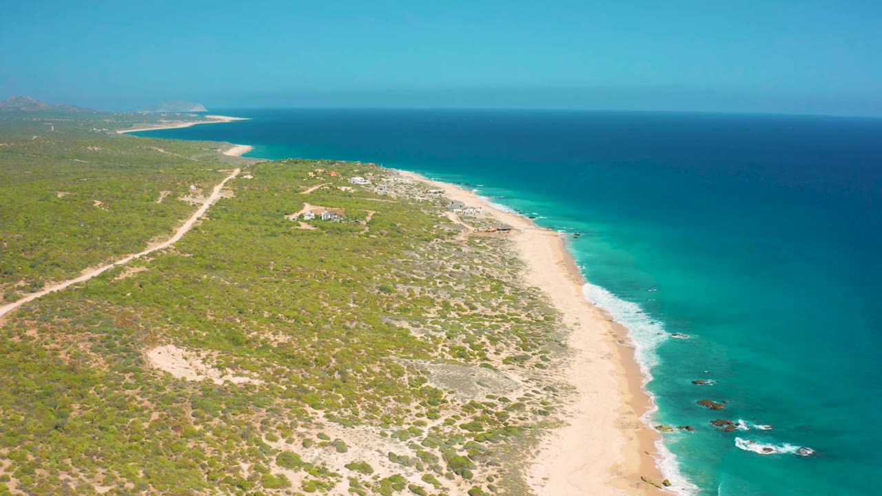 High aerial frame at El Cardoncito showing a diagonal strip of pale sand, low vegetation, dirt tracks inland, turquoise shallows, and no visible people.