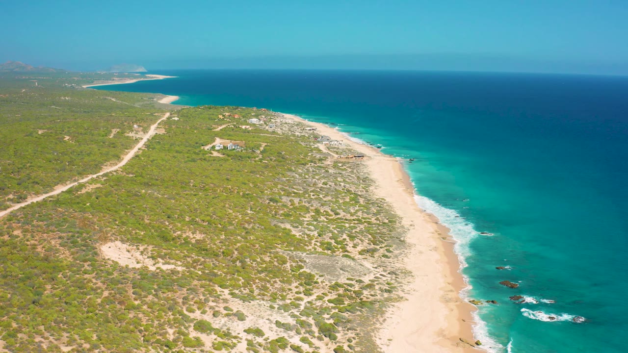 Drone-style aerial over El Cardoncito with coastal scrub in the foreground, a bright sand beach along the waterline, rocks in the surf, and no visible people.