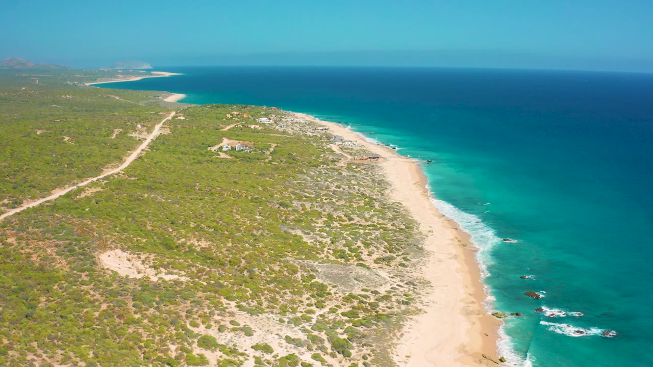Aerial view at El Cardoncito with inland dirt roads crossing low vegetation, a narrow beach edge, small waves breaking along shore, and no visible people.