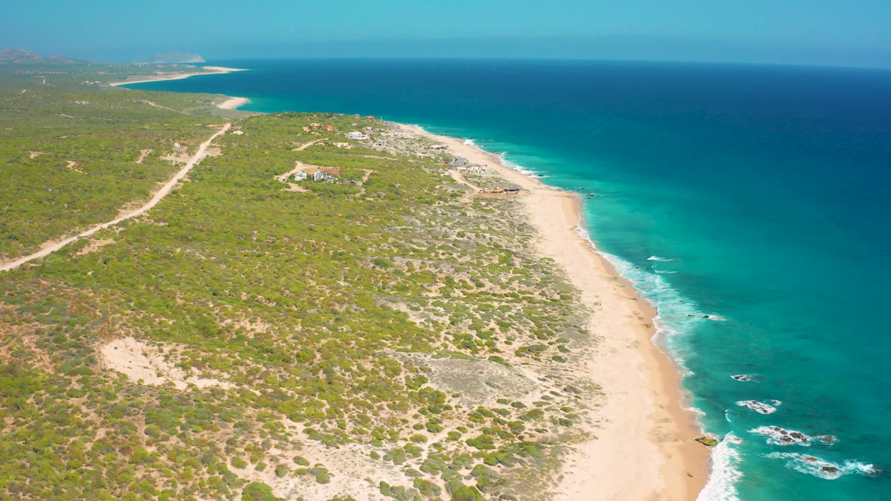Aerial frame over El Cardoncito, Baja California Sur, showing a diagonal sandy beach, turquoise water, low green bushes, inland dirt road, and no visible people.