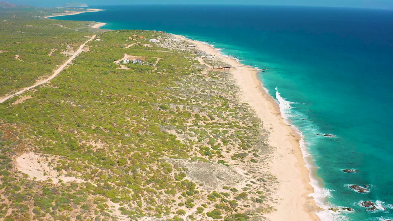 Aerial frame at El Cardoncito with the sandy shoreline slightly farther along the coast, white surf on the right, scattered rocks in the water, inland scrub, and no visible people.