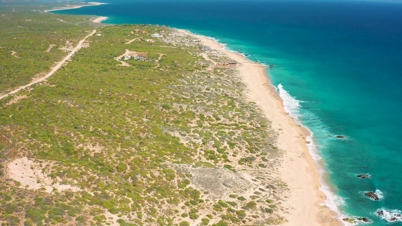 Aerial view over Baja California Sur coastline with more nearshore rocks visible in the lower right surf, beach running diagonally, inland dirt tracks, scrub, and no visible people.