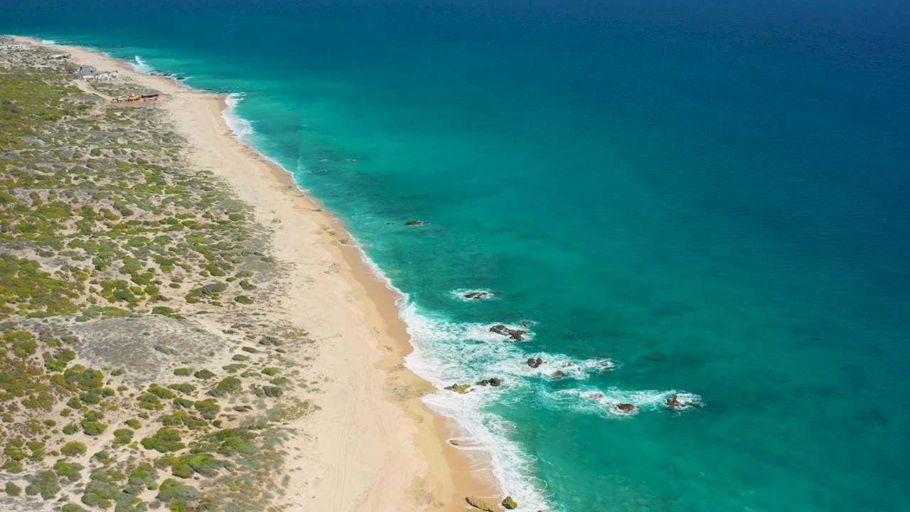 Aerial view of sandy beach, scrubland, turquoise water, and small rocks in the surf.