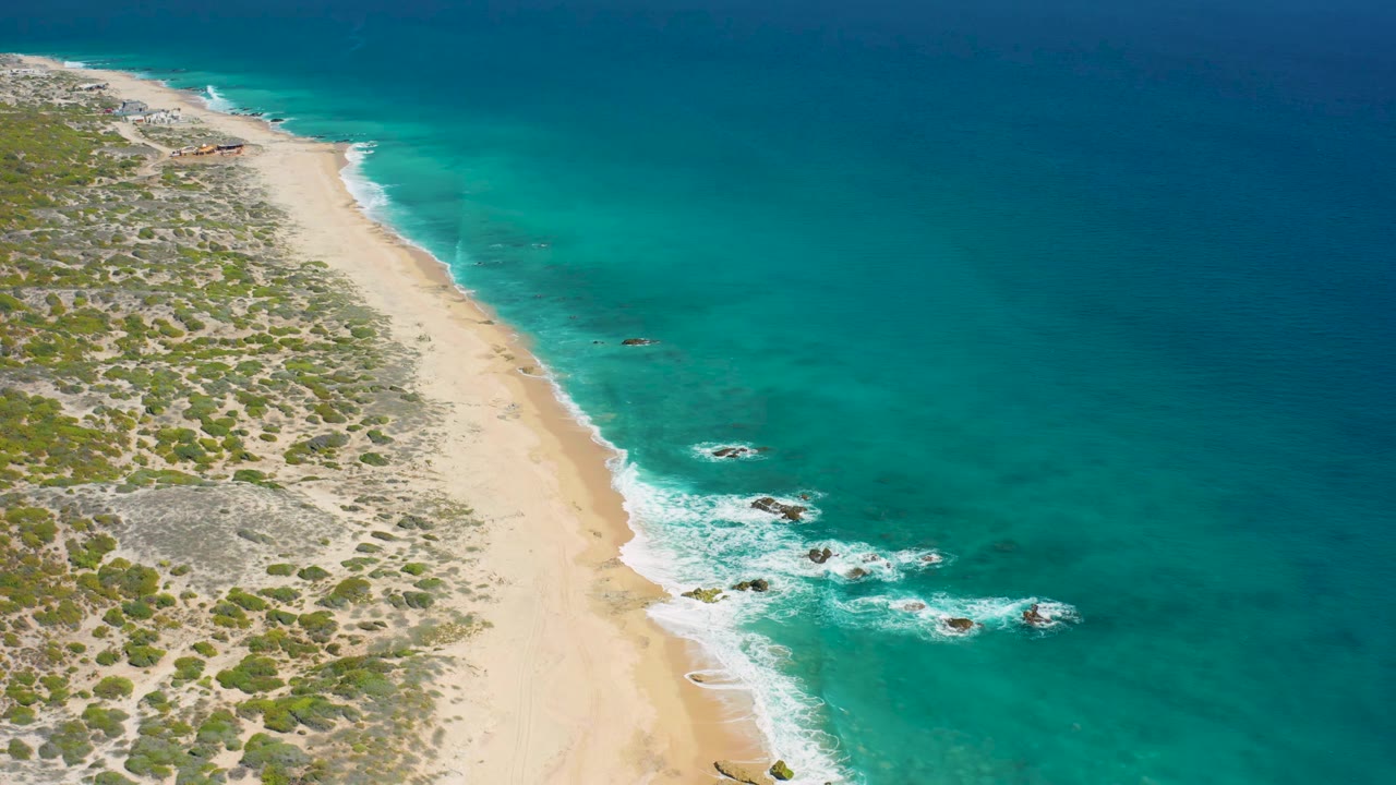 Aerial view of El Cardoncito shoreline as white surf curls around dark rocks near the beach, with scrubland on the left and no visible people.