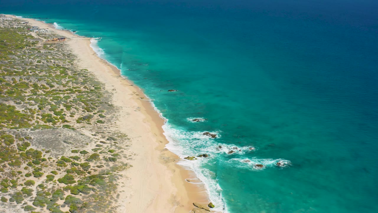 El Cardoncito beach seen from above with brighter turquoise water close to shore, darker ocean beyond, scattered rocks in the surf, and no visible people.