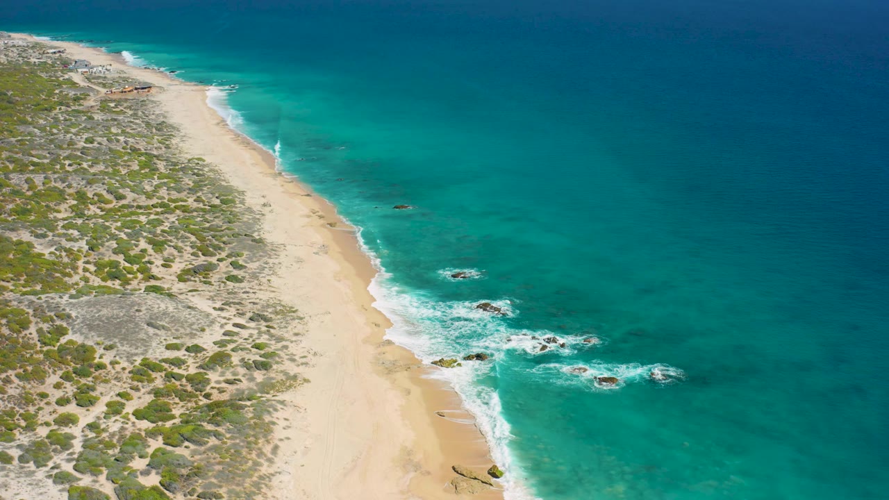 Overhead coastal view at El Cardoncito with the beach centered between scrubland and turquoise sea, while small white waves mark the shore and no visible people appear.
