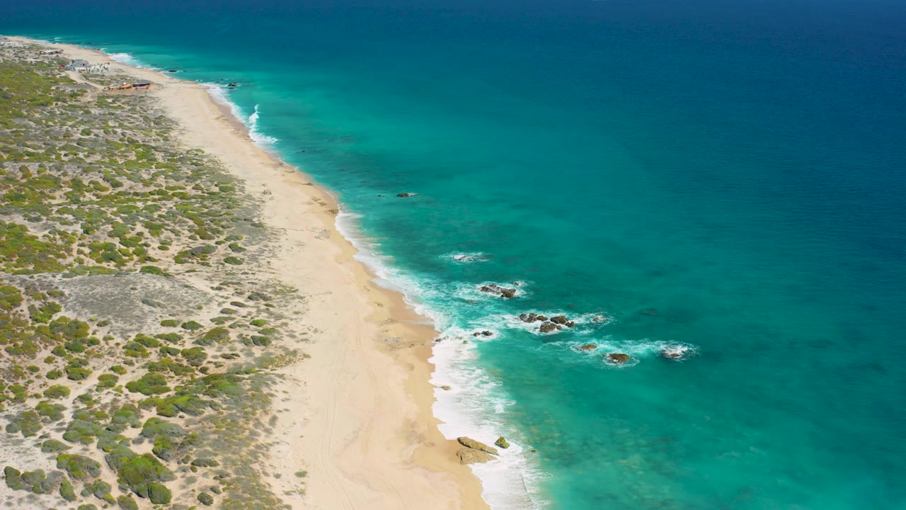 Overhead view of El Cardoncito coastline with pale sand, low scrub vegetation, turquoise water, scattered rocks in the surf, and no visible people.