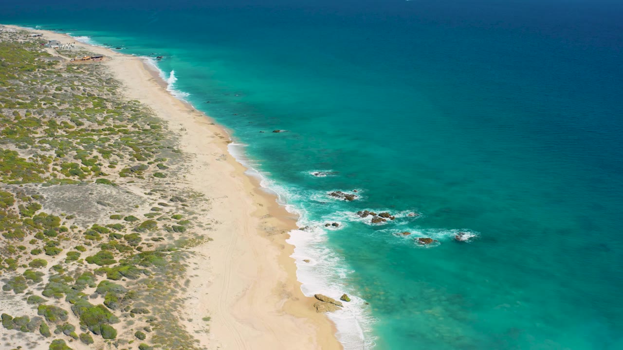 El Cardoncito shoreline from above with a broad empty sand strip, turquoise sea to the right, small nearshore rocks, white wave lines, and no visible people.