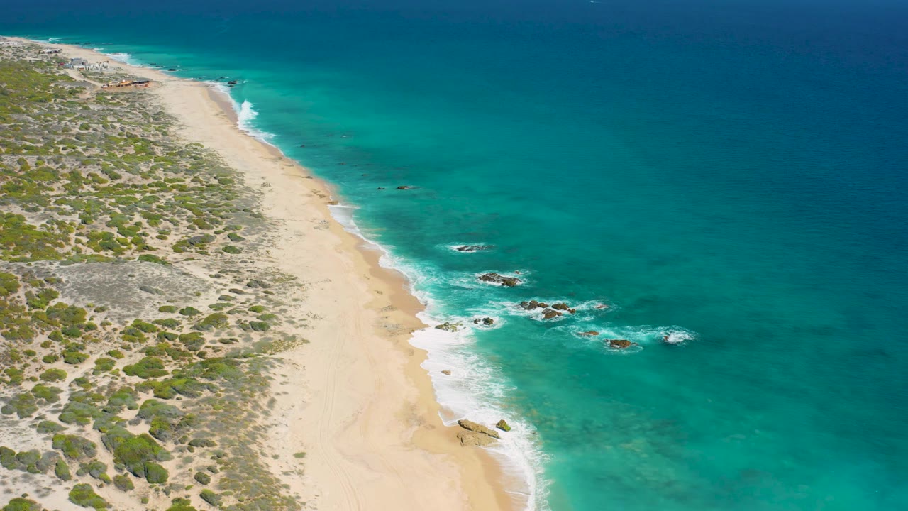 Aerial view of El Cardoncito with more scrub terrain visible along the left edge, a pale beach corridor, turquoise shallows, surf foam, and no visible people.