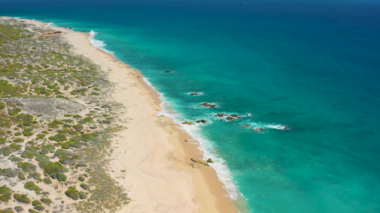 Opening aerial frame over El Cardoncito, Baja California Sur, with scrub-covered dunes on the left, pale beach below, turquoise water on the right, small rocks offshore, and no visible people.