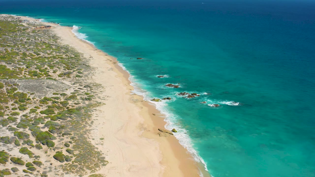 El Cardoncito aerial frame with empty sand in the lower left, clear shallow water near shore, darker ocean farther out, small rock clusters with white surf, and no visible people.
