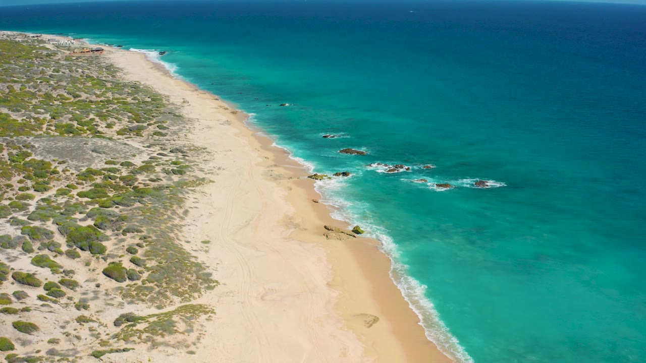Opening aerial frame over El Cardoncito beach with pale sand, low scrubland on the left, turquoise shallows on the right, scattered offshore rocks, and no visible people.