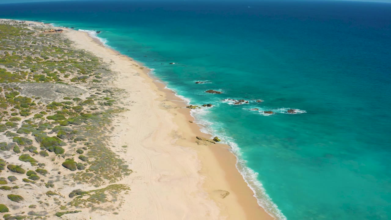 Aerial view along the El Cardoncito shoreline with the beach running diagonally through frame, scrub-covered dune terrain inland, aqua surf along the waterline, and no visible people.