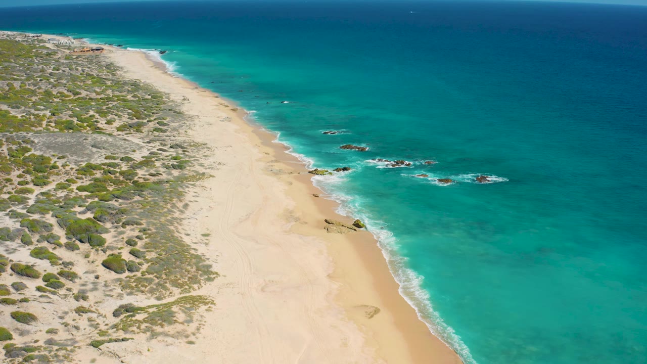 Wide elevated frame over El Cardoncito with a long pale beach, blue-green water bands, small rocks beyond the surf line, low vegetation inland, and no visible people.