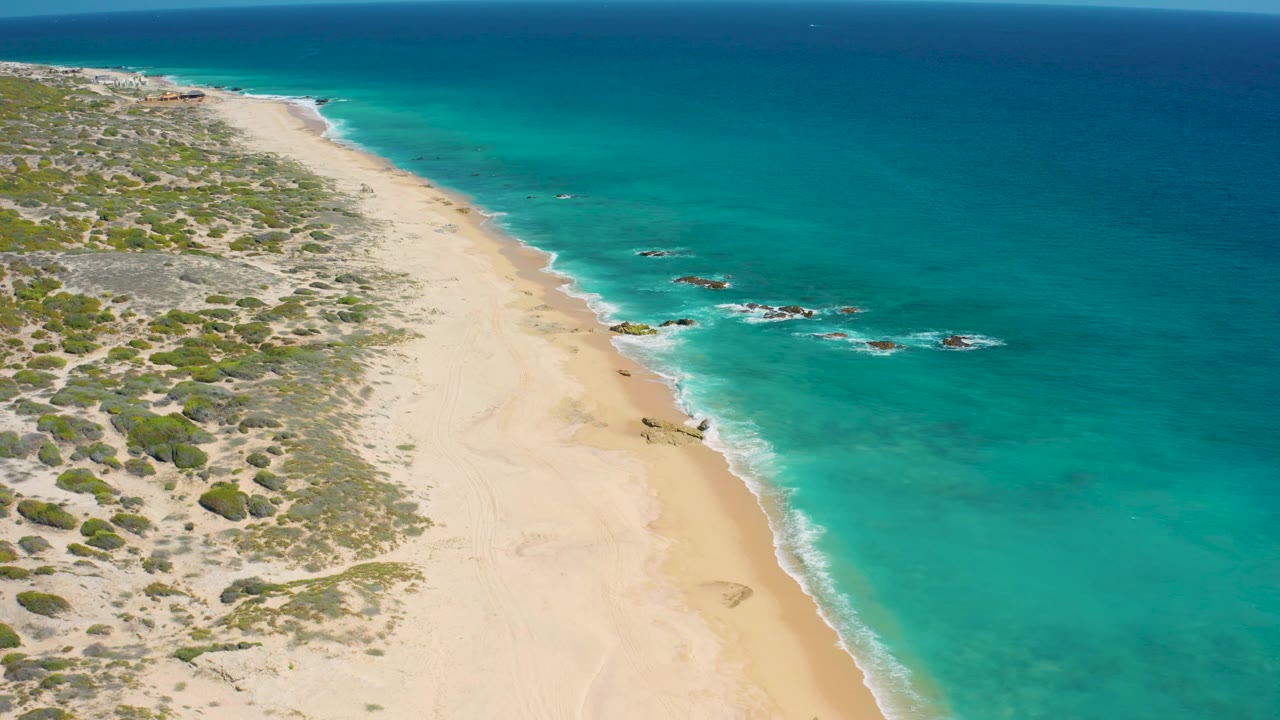 Aerial frame looking down the El Cardoncito coast as the empty sand curves beside calm turquoise sea, with scrub and dune textures filling the inland side and no visible people.