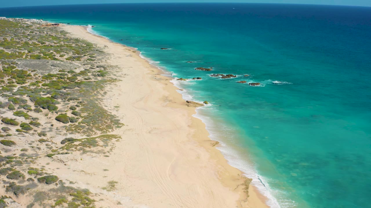 Wide drone-like frame of El Cardoncito shoreline, where small rock outcrops sit beyond the wave line and the empty beach angles toward the horizon.