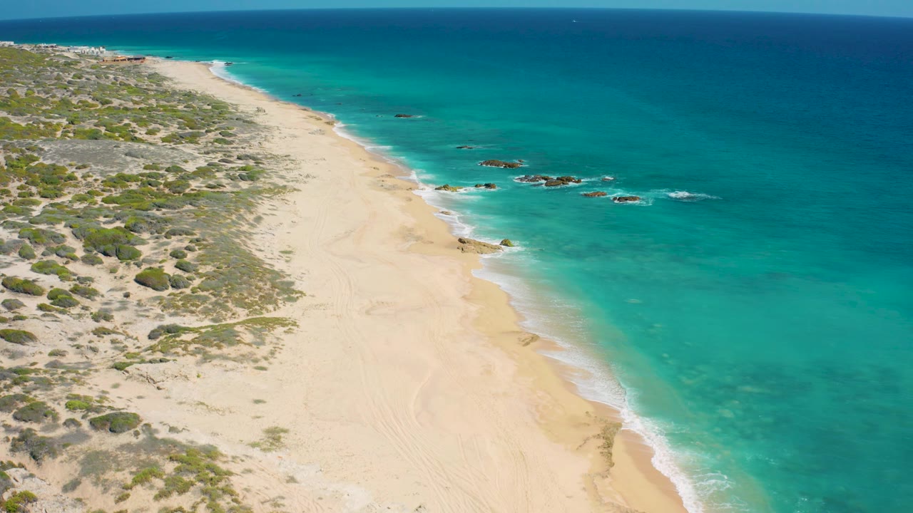 Aerial view along the empty El Cardoncito beach with bright turquoise water on the right, scrub-covered dunes on the left, and no visible people.