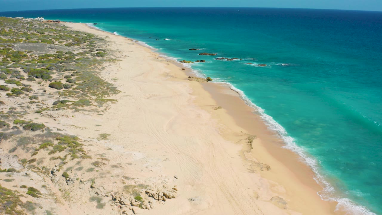 Aerial frame over El Cardoncito showing pale sand, low coastal scrub, rocky patches, turquoise nearshore water, and no visible people.