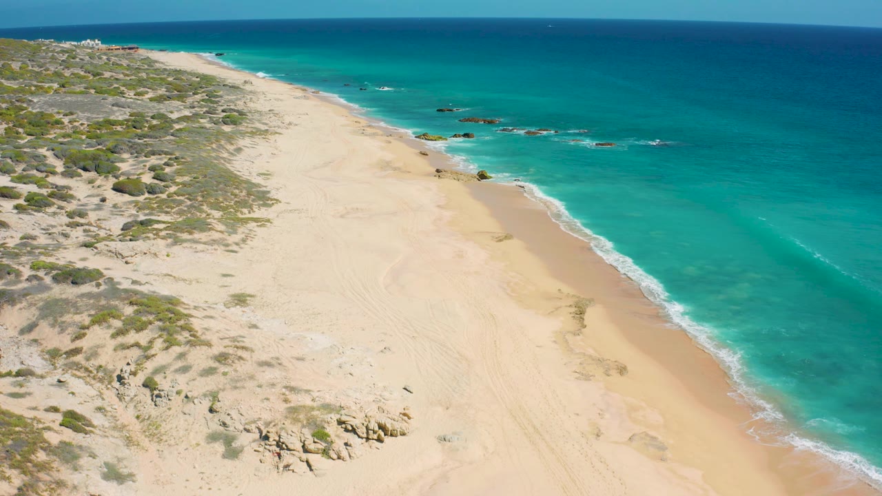 Aerial frame over El Cardoncito with a broad empty beach, scattered rocks near the lower center, turquoise shallows, offshore rock outcrops, and no visible people.