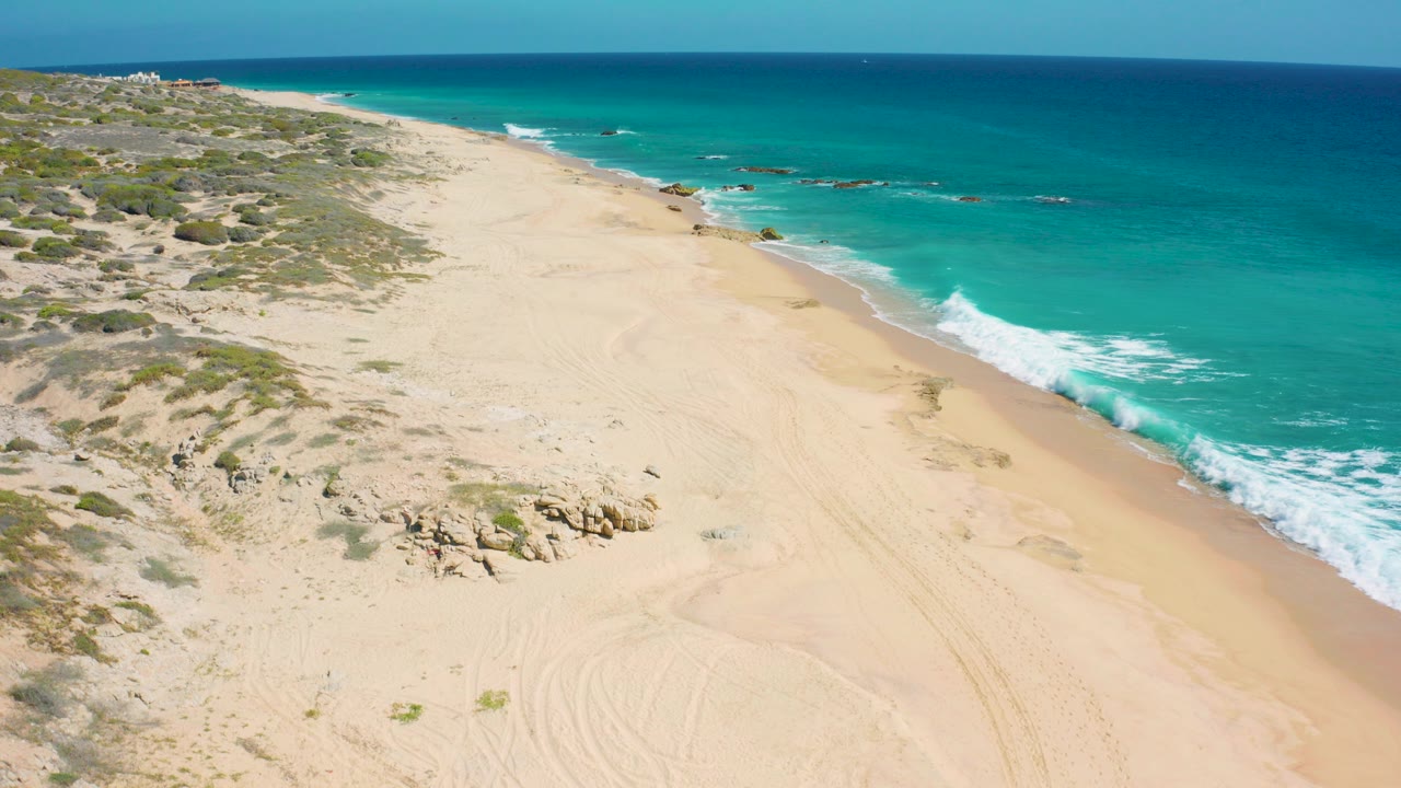 Elevated view over El Cardoncito beach in Baja California Sur, with pale sand, low green scrub, rocks, turquoise water, white surf, and no visible people.
