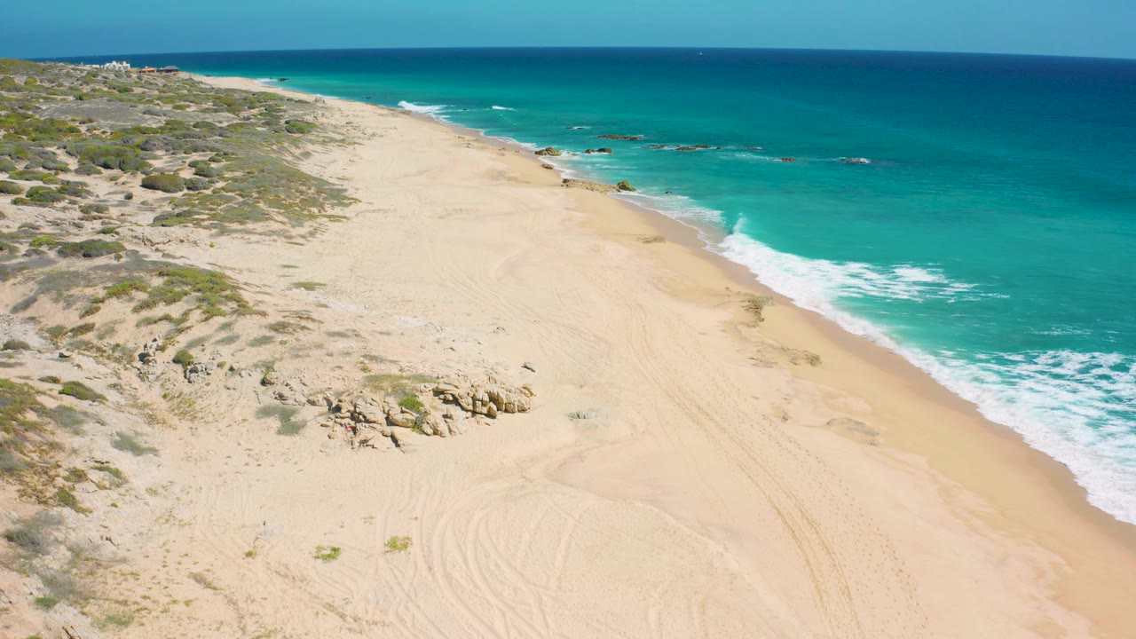 El Cardoncito coastline seen from above, with broad pale sand in the center, low brush and rocks to the left, blue-green sea to the right, and no visible people.