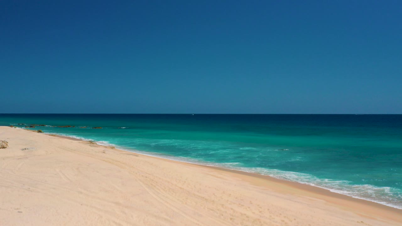 Opening frame over El Cardoncito, Baja California Sur, showing pale sand at left, turquoise water along the shore, deep blue sea beyond, clear sky, and no visible people.