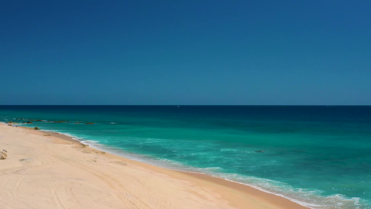The view over El Cardoncito shows less sand and more water, with small dark patches offshore, a thin surf line, open sea, clear sky, and no visible people.