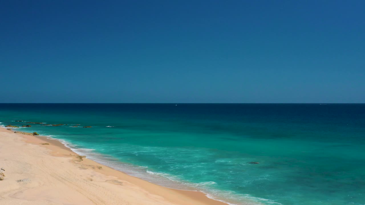 Mid-clip frame at El Cardoncito with a broad sky band, straight sea horizon, turquoise water across the middle, beach entering from the lower left, and no visible people.